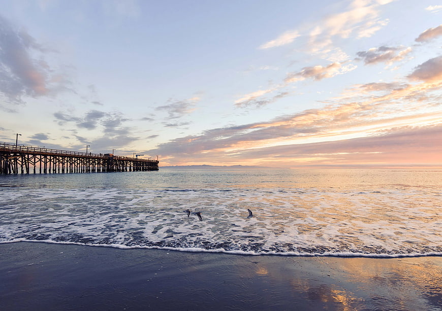 Sunset over ocean pier with seagulls flying low over waves