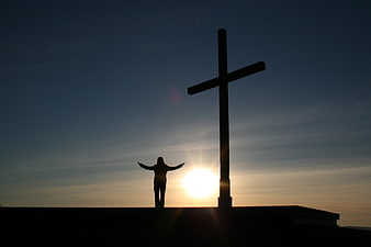 Silhouette of person with arms raised near cross at sunset