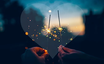 Hands holding heart cutout with sparklers against sunset