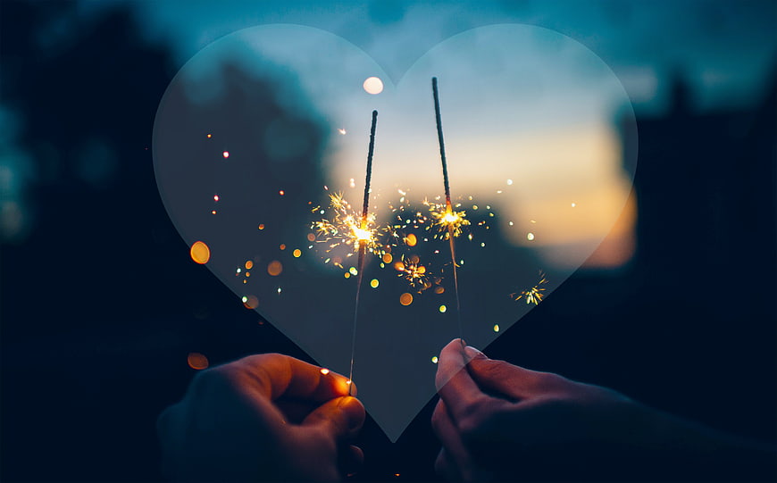 Hands holding heart cutout with sparklers against sunset