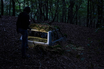 Person standing by abandoned mossy car in dark forest