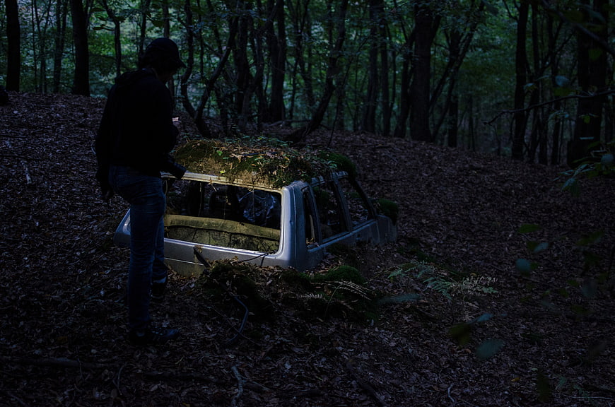 Person standing by abandoned mossy car in dark forest