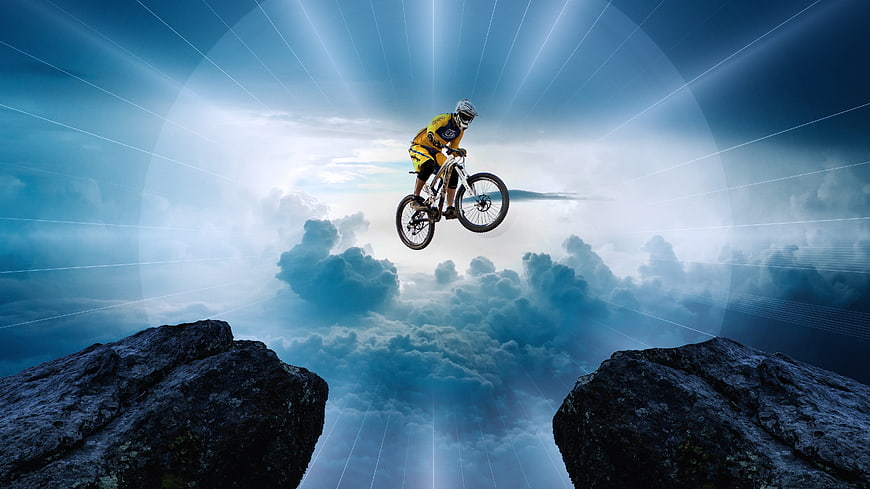 Mountain biker jumping between cliffs above clouds with light rays