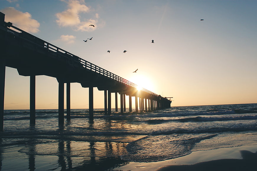 Silhouette of pier at sunset with birds flying over ocean waves