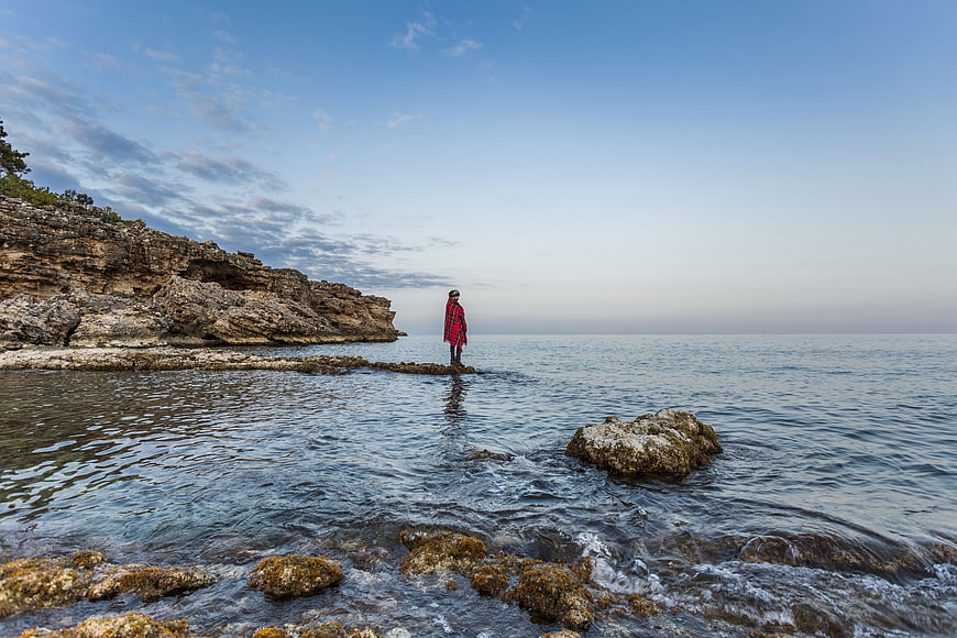 Man in Red Coat Standing on Rocky Shoreline at Dusk